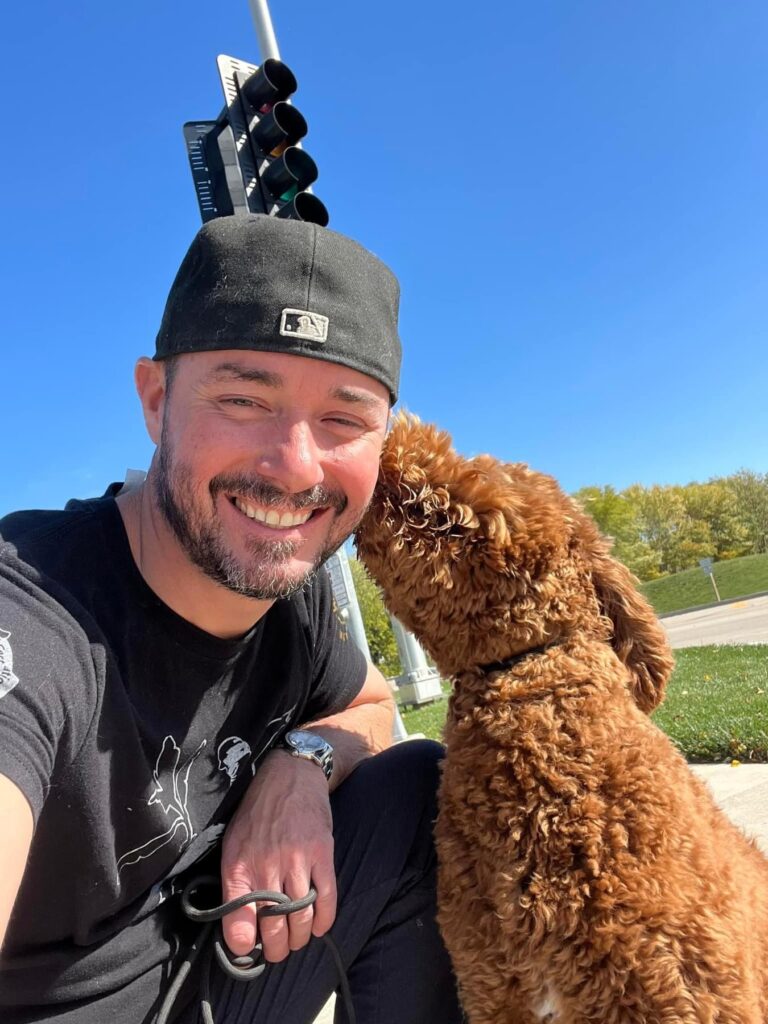 Tyler Morrick, Head Trainer with TTl on a walk with a golden doodle. The dog is a deep brown and giving him a kiss.