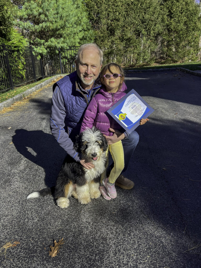 A Bernedoodle posing outdoors with its owner holding a certificate after completing the board and train program