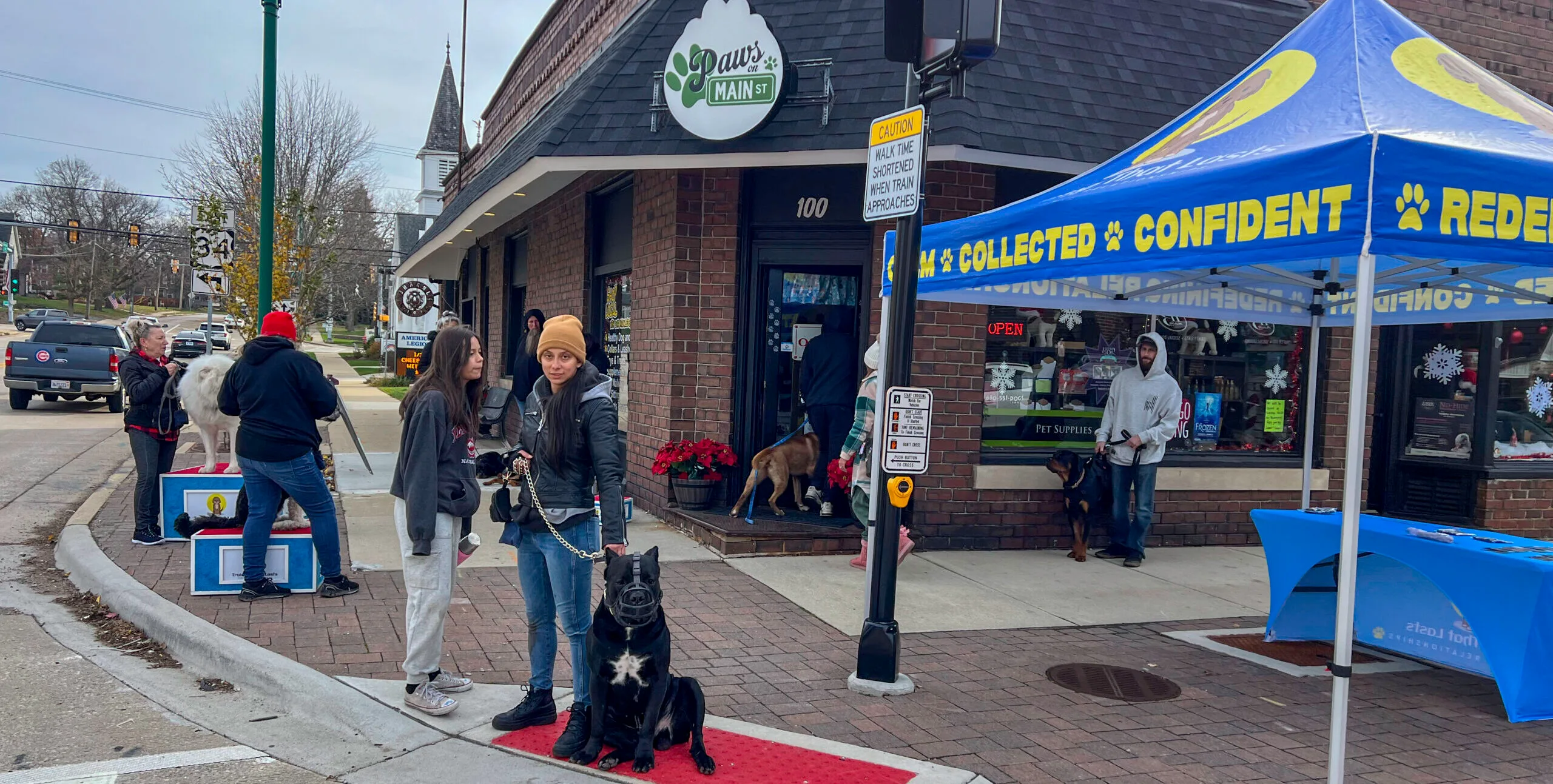 A Group Of People Standing Next To Their Dogs In Front Of Paws On Main St In Oswego During Training That Lasts Group Training Session.