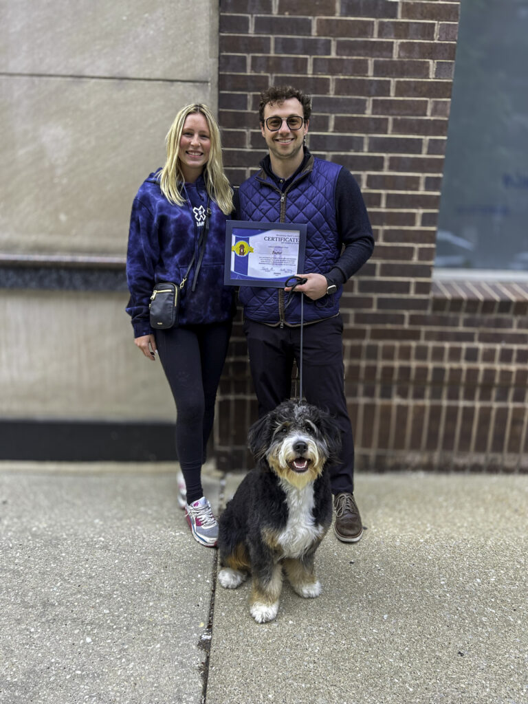 A Bernedoodle standing next to its owner outside a building, showcasing their board and train program certificate