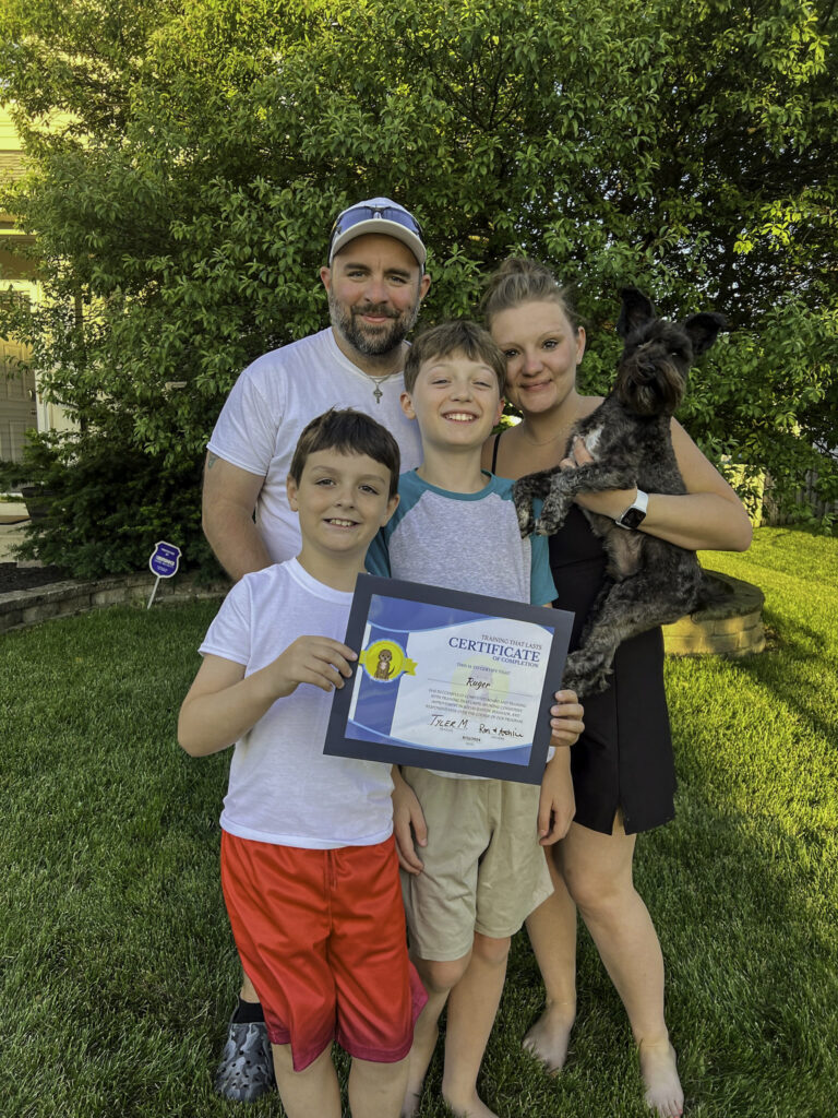 A family of four smiling with their Bernedoodle holding its board and train graduation certificate outdoors