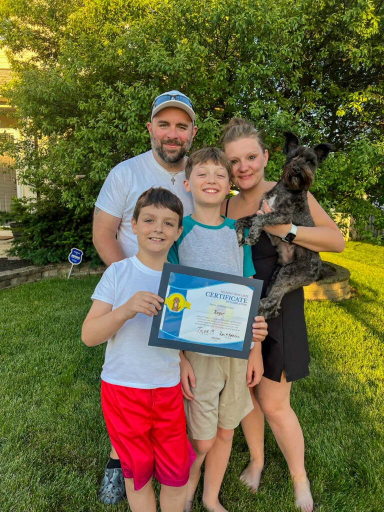 A Family Of Four Smiling With Their Bernedoodle Holding Its Board And Train Graduation Certificate Outdoors